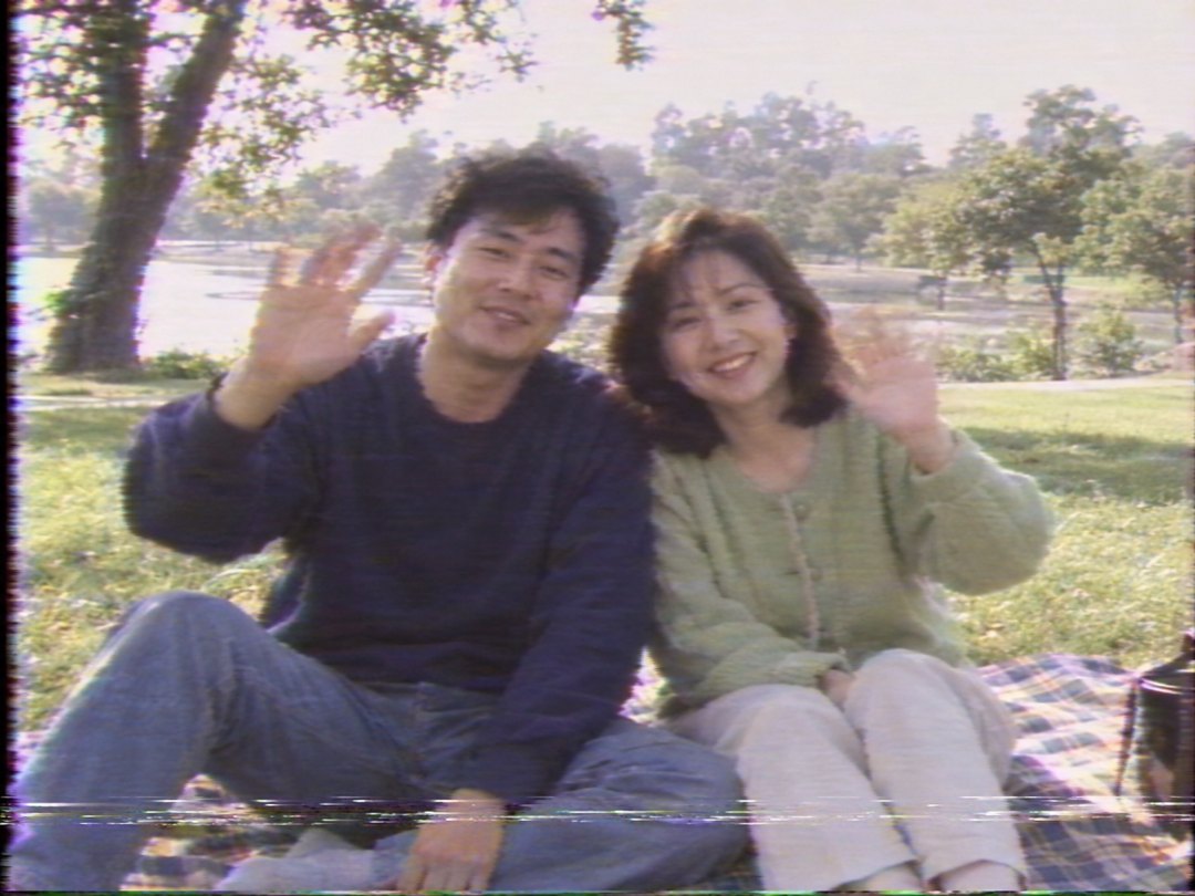 지어낸 30대 부부가 공원 돗자리에 앉아 손을 흔드는 장면을 4:3 화면, 흐리고 번진 색, 아래쪽 가로 줄무늬 잡음이 있는 VHS 홈비디오 정지 화면처럼 그린 그림